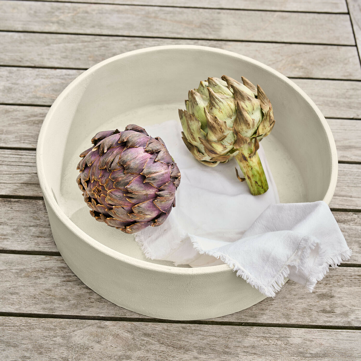 Centrotavola Islene Ciotola rotonda in ceramica color crema con due carciofi e panno bianco su tavolo in legno
