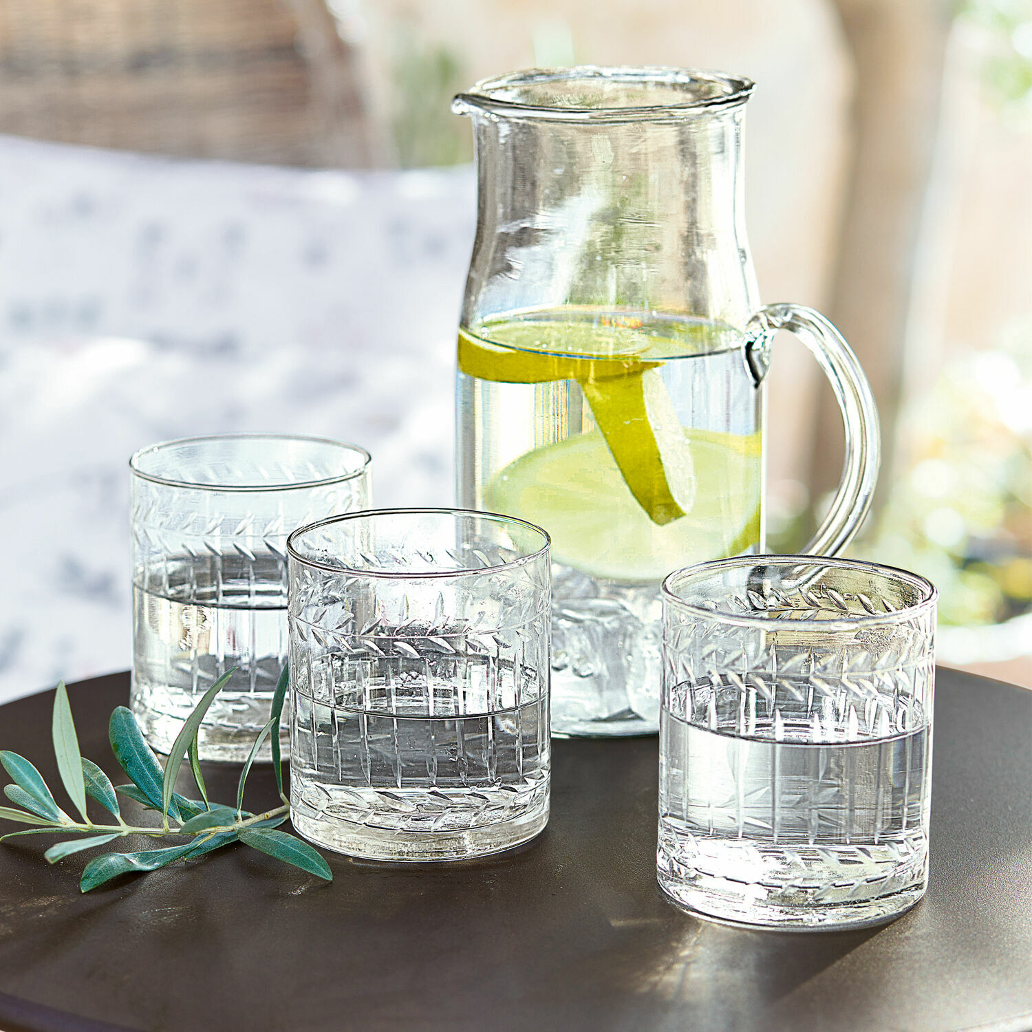 Set di 6 bicchieri Padiès Caraffa di vetro con fette di limone e tre bicchieri d'acqua su tavolo scuro con foglie verdi decorative