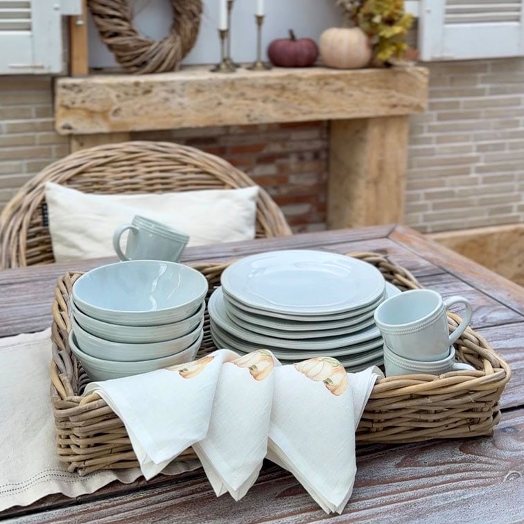 Set di stoviglie in ceramica grigio chiaro con piatti, ciotole e tazze in un cesto di vimini su tavolo in legno