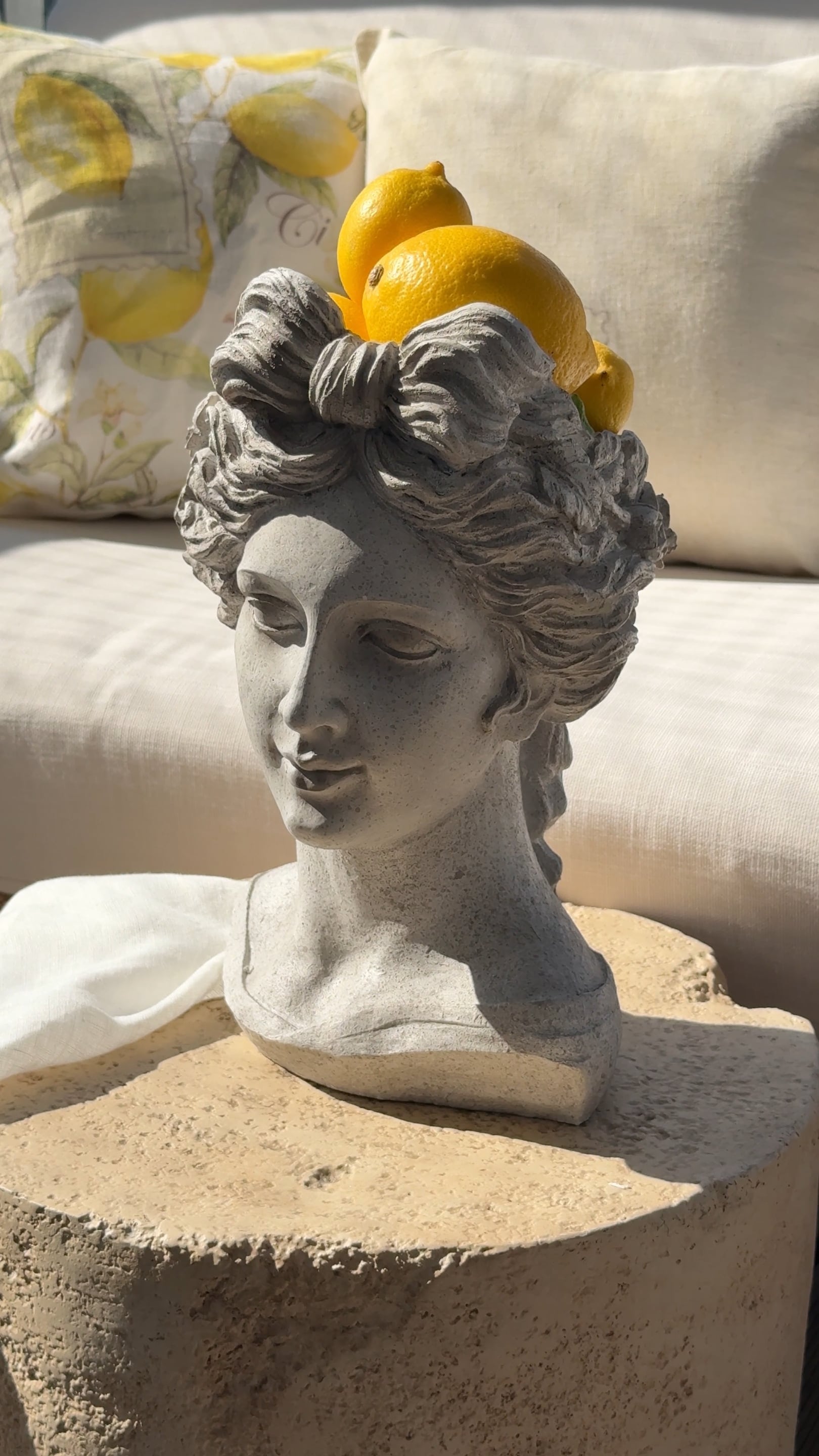 Busto in pietra grigia con limoni sulla testa davanti a cuscini chiari e tessuti beige