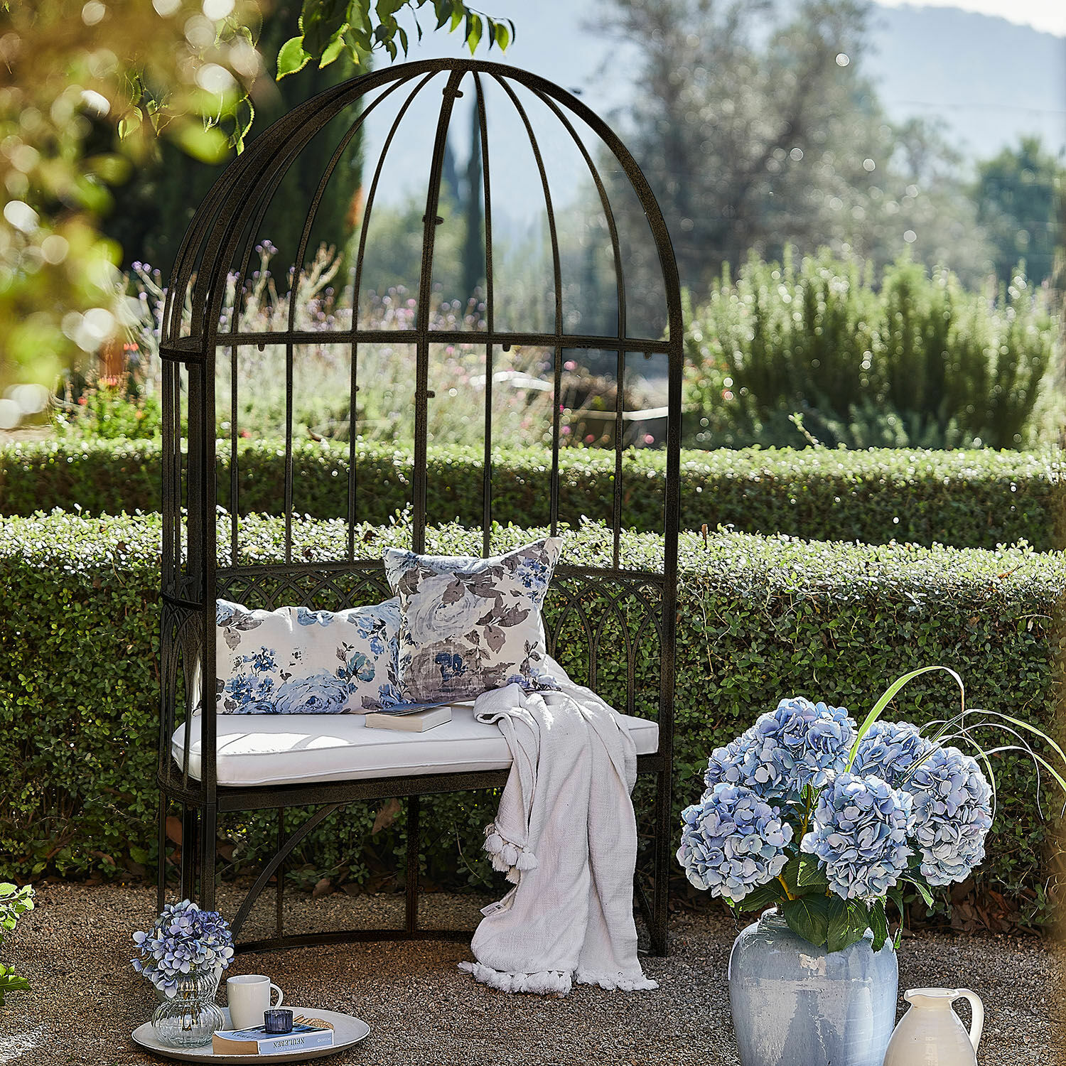Panchina Rivandelle Giardino con panchina in metallo a forma di gabbia, cuscino bianco e fiori blu in vaso