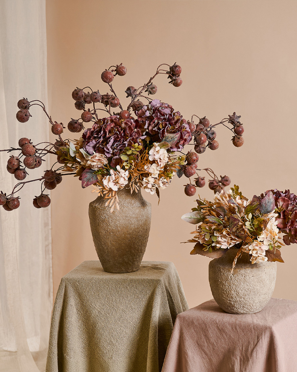 Due vasi decorativi in pietra beige con fiori autunnali e frutti secchi su tavoli coperti