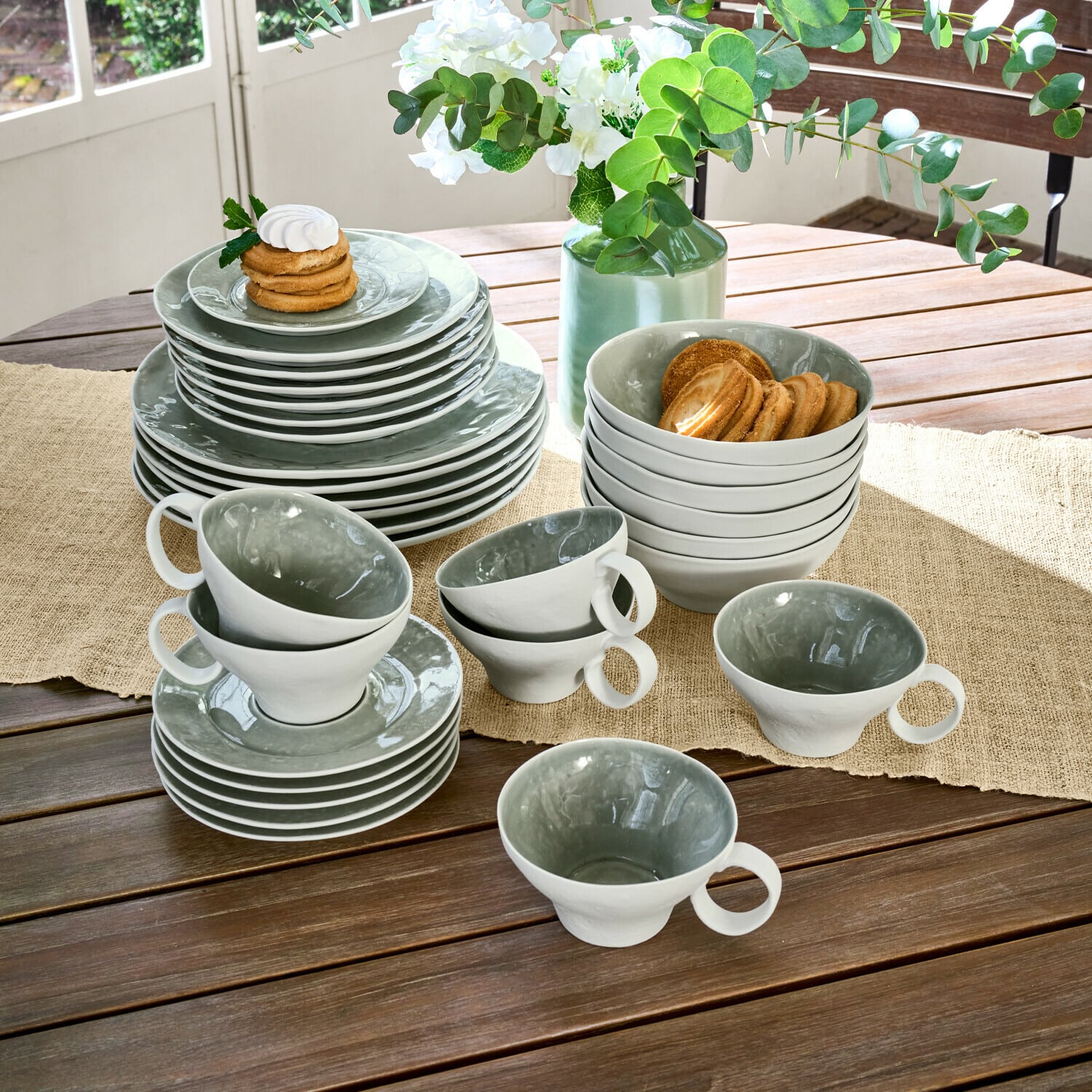 Set di 30 stoviglie Braganza Set di stoviglie in ceramica grigia e bianca su tavolo in legno con biscotti e fiori decorativi
