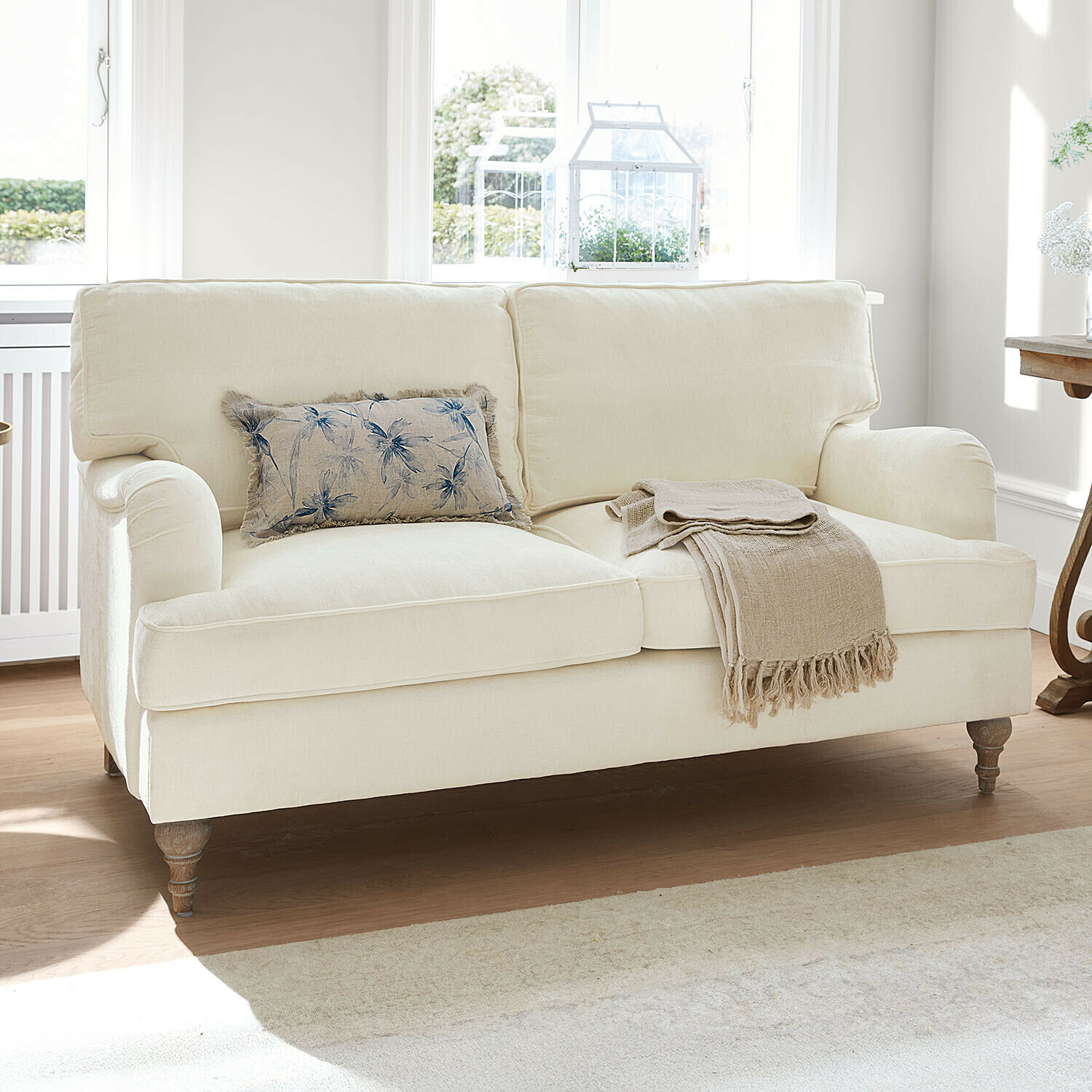 Divano Elvika Divano crema imbottito con cuscino floreale e coperta beige in soggiorno luminoso