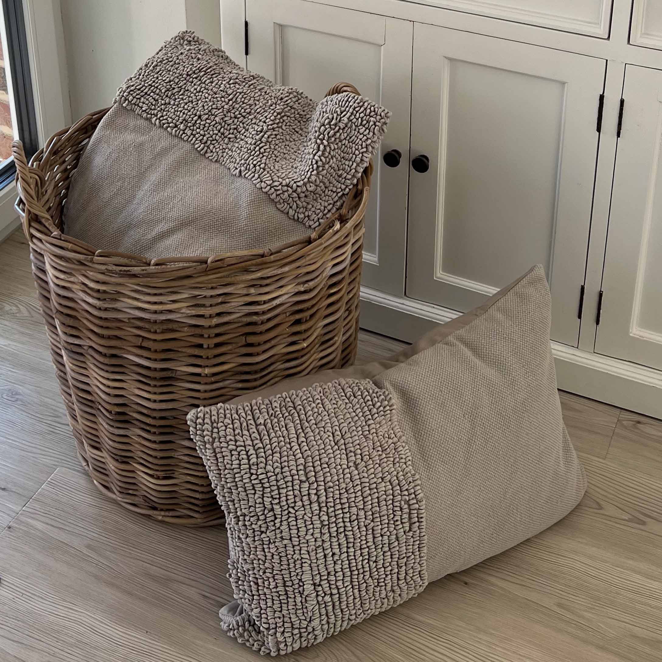 Cuscino beige in materiale morbido con texture accanto a un cesto di vimini su pavimento in legno