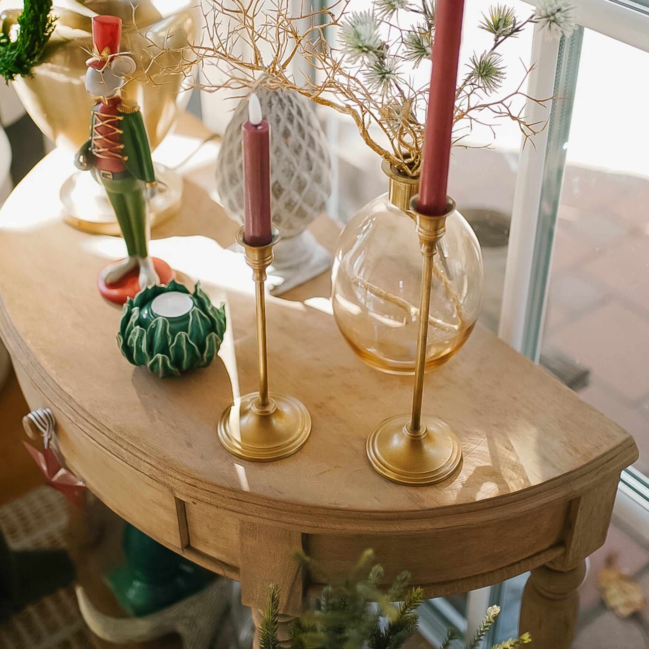 Tavolo in legno con decorazioni natalizie, candelabri dorati e candele rosse vicino a una finestra