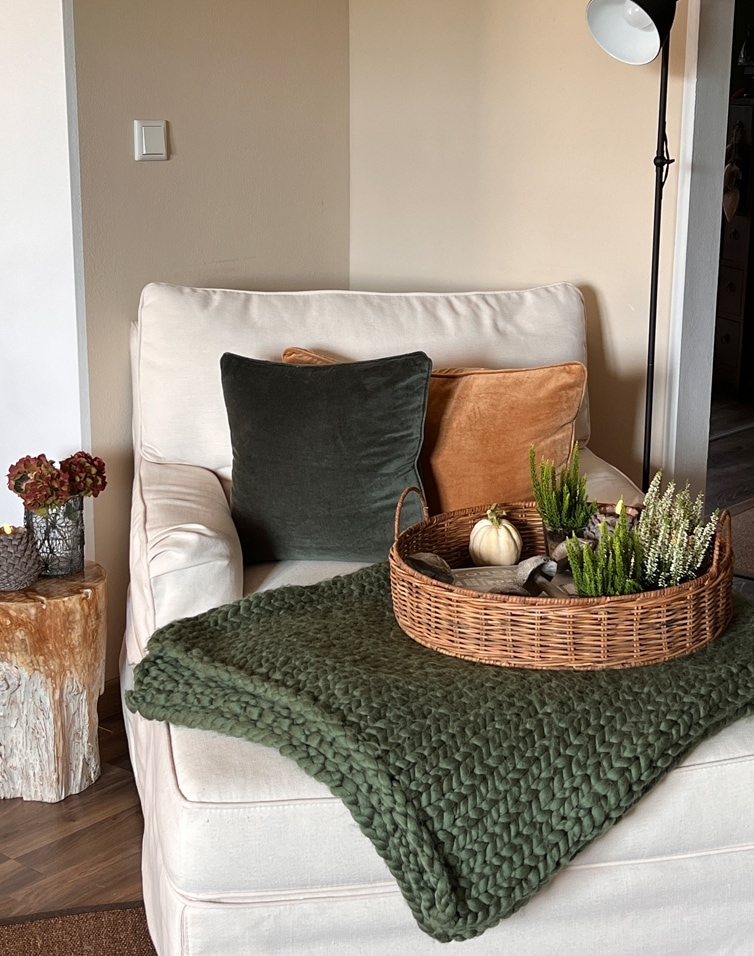 Poltrona beige con cuscini verdi e arancioni, cesta di vimini con decorazioni su coperta verde