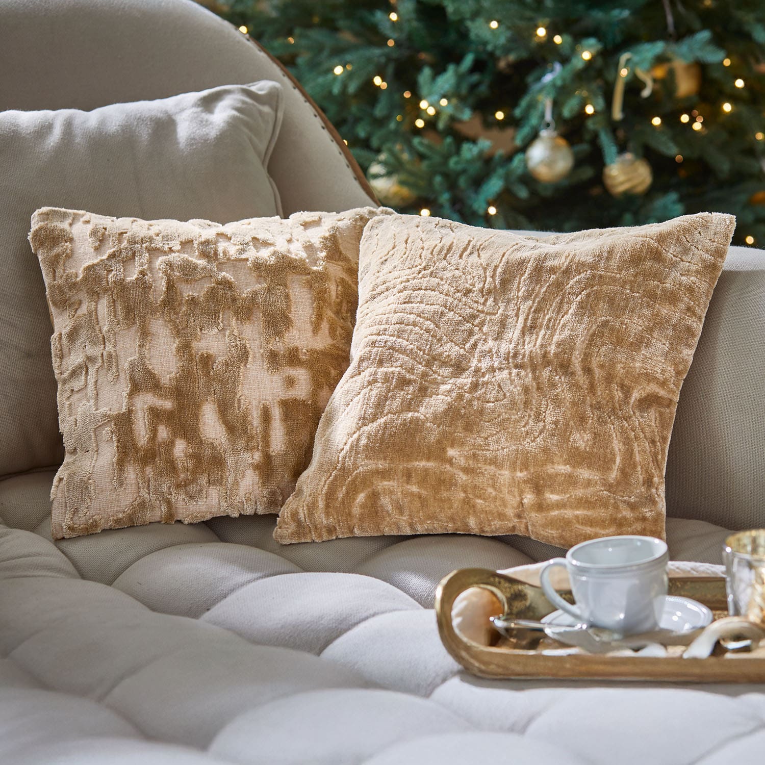 Due cuscini decorativi beige in tessuto morbido su divano chiaro vicino a un albero di Natale decorato