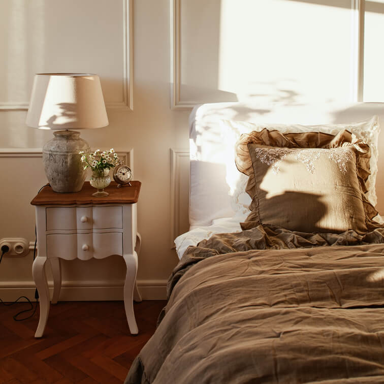 Camera accogliente con biancheria da letto beige e comodino bianco con lampada e fiori