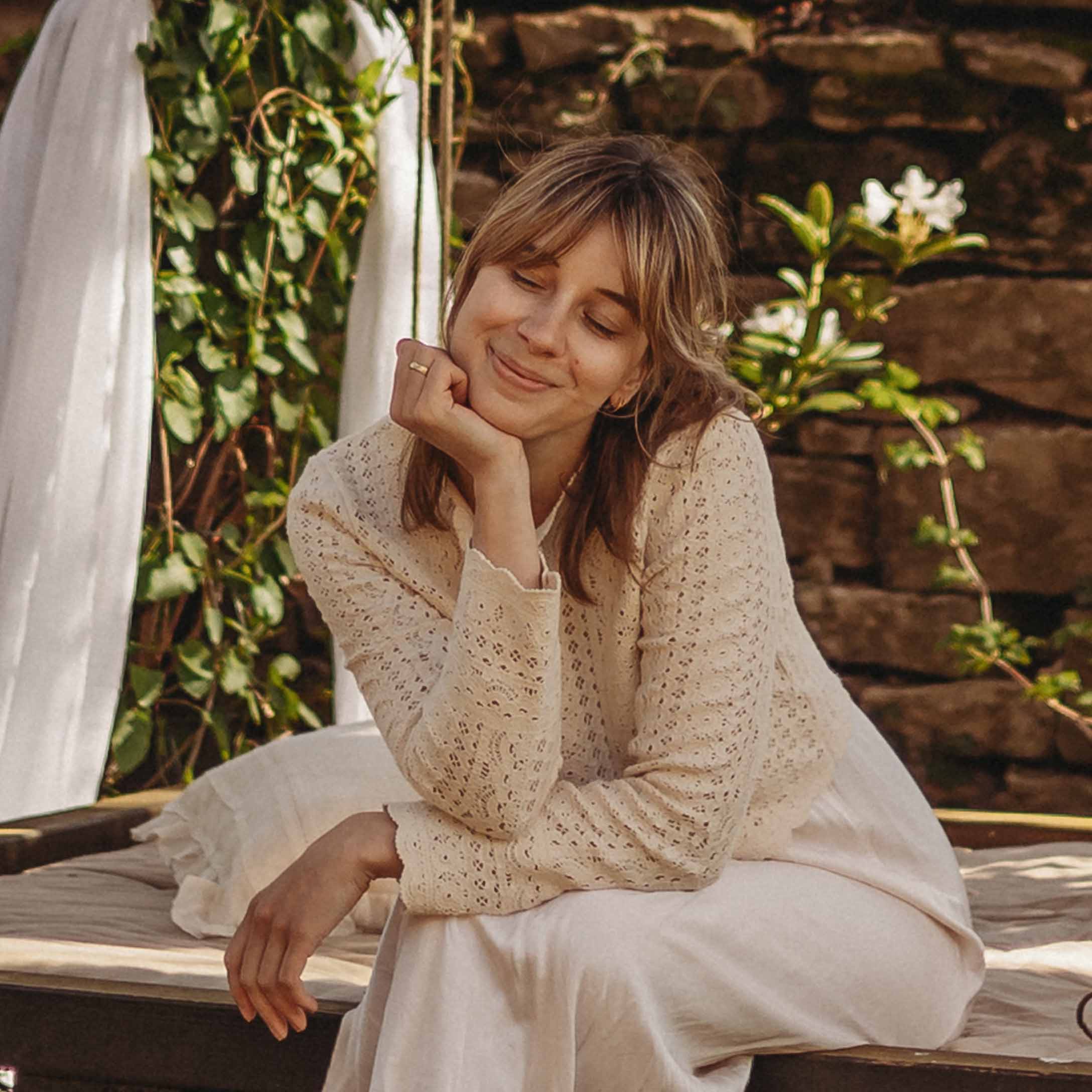 Donna con maglione traforato crema seduta all'aperto su cuscino in ambiente naturale