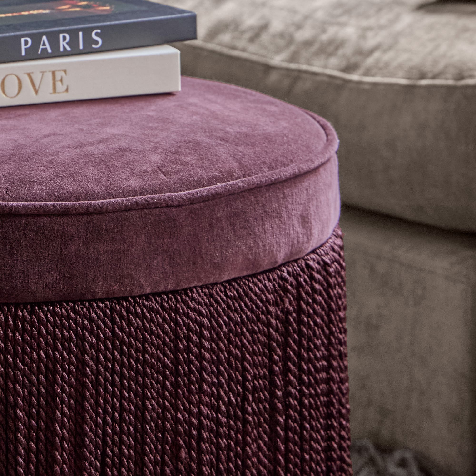 Pouf viola in velluto morbido e tessuto intrecciato con divano grigio e libri sullo sfondo