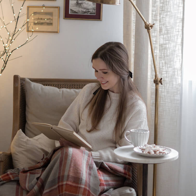 Donna che legge un libro su poltrona in vimini con maglione crema e coperta calda a quadri