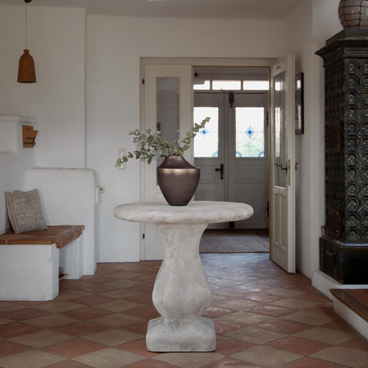 Tavolo rotondo in cemento grigio chiaro con vaso in ceramica scura e foglie verdi in interno luminoso