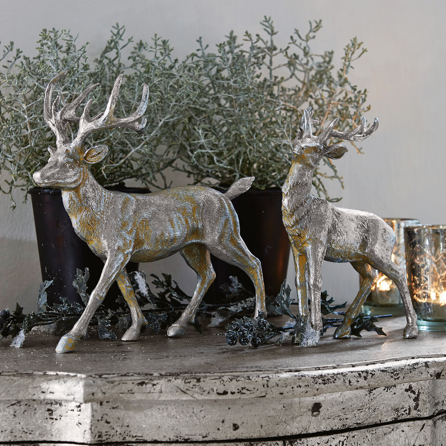 Coppia di cervi Rudy & Son Due decorazioni di cervi argentati in resina su una credenza rustica con piante e candele