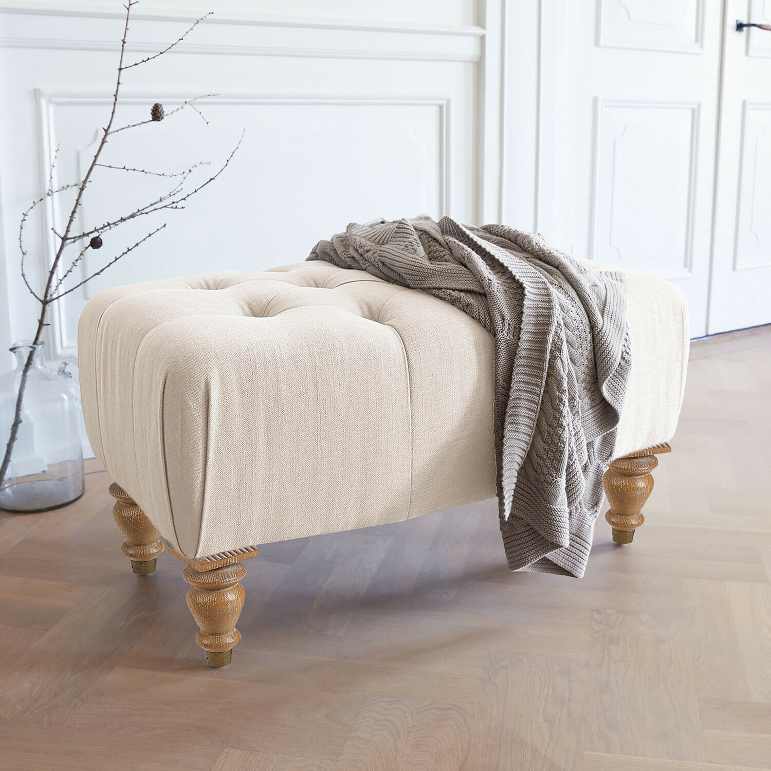 Pouf Durance Pouf beige imbottito con gambe in legno e coperta lavorata a maglia grigia in interno luminoso