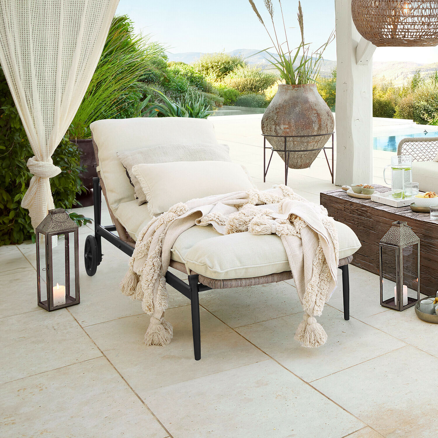 Lettino da giardino con cuscini crema e coperta morbida su terrazza con vista piscina e verde