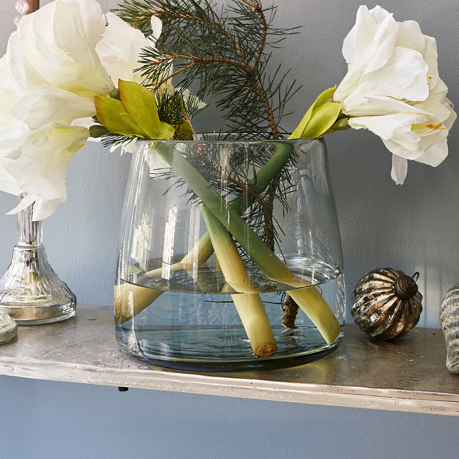 Vaso Inga Vaso di vetro trasparente con fiori bianchi e rami verdi su una mensola decorativa
