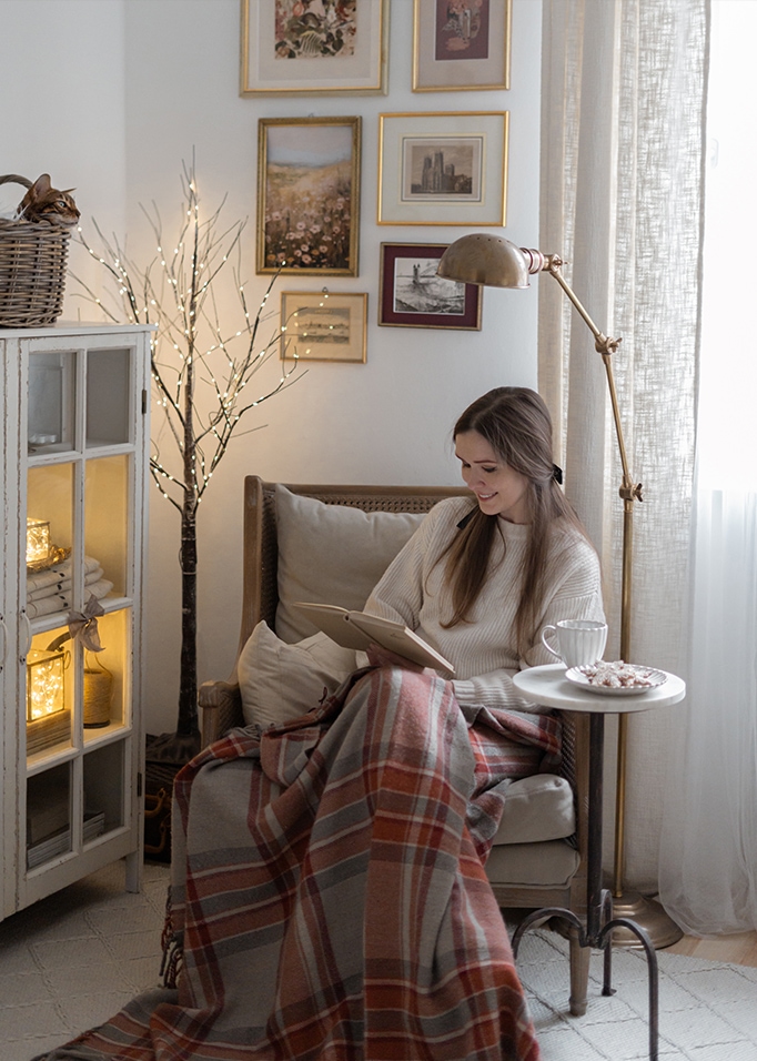 Donna che legge un libro sotto una coperta calda in un soggiorno accogliente con lampada e decorazioni murali