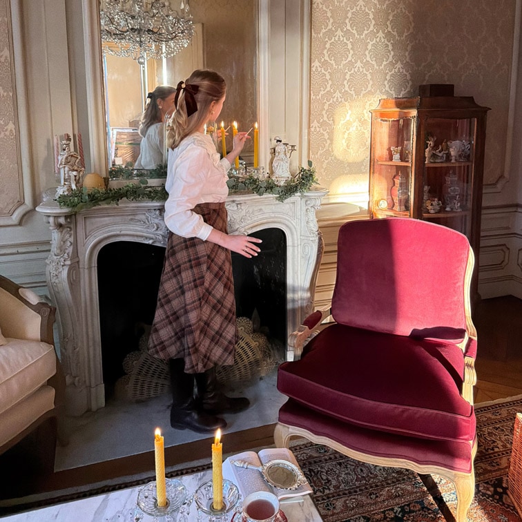 Donna che accende candele vicino a un camino in marmo in un salotto elegante con poltrona bordeaux