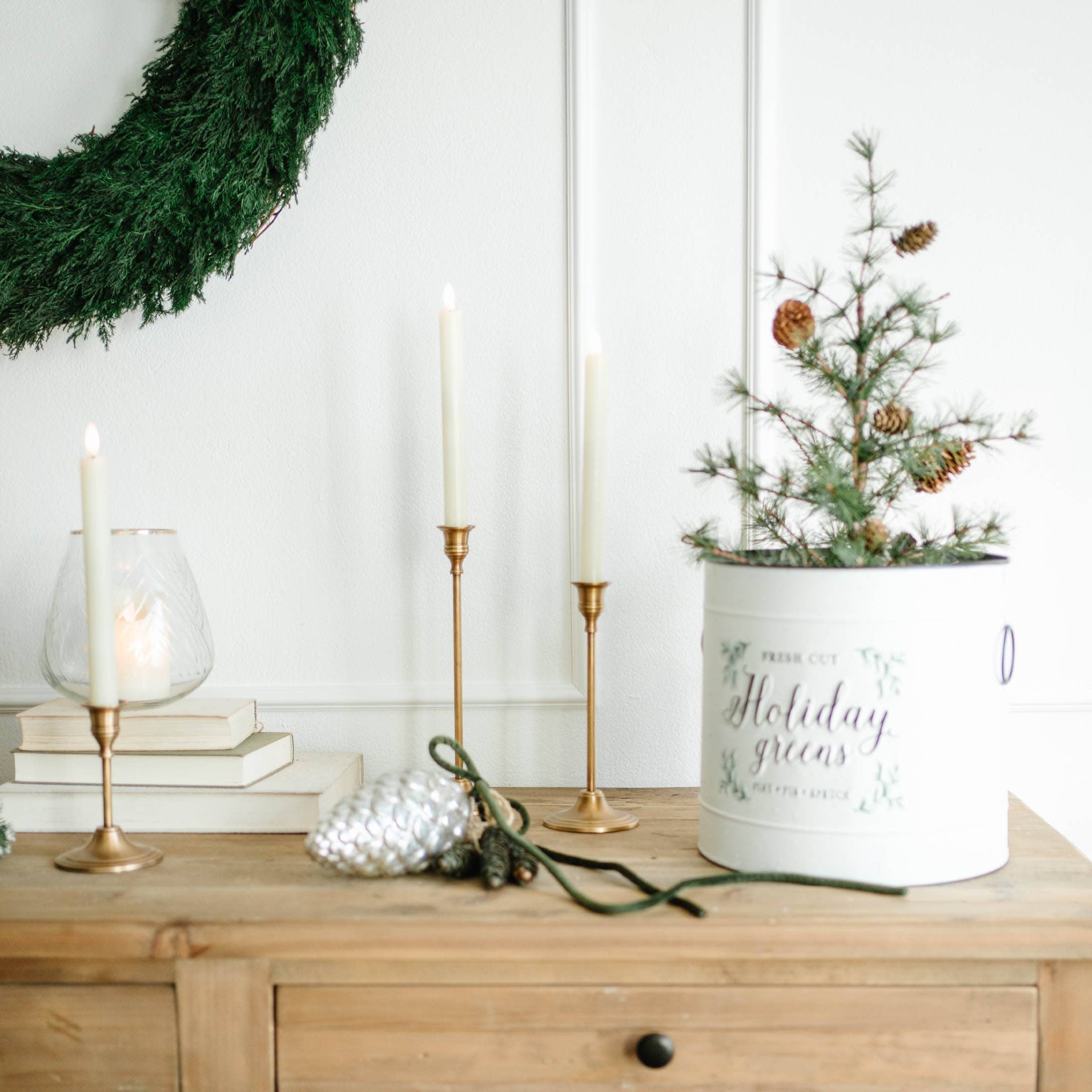 Decorazione natalizia con corona verde, candelabri dorati e contenitore bianco con rami e pigne