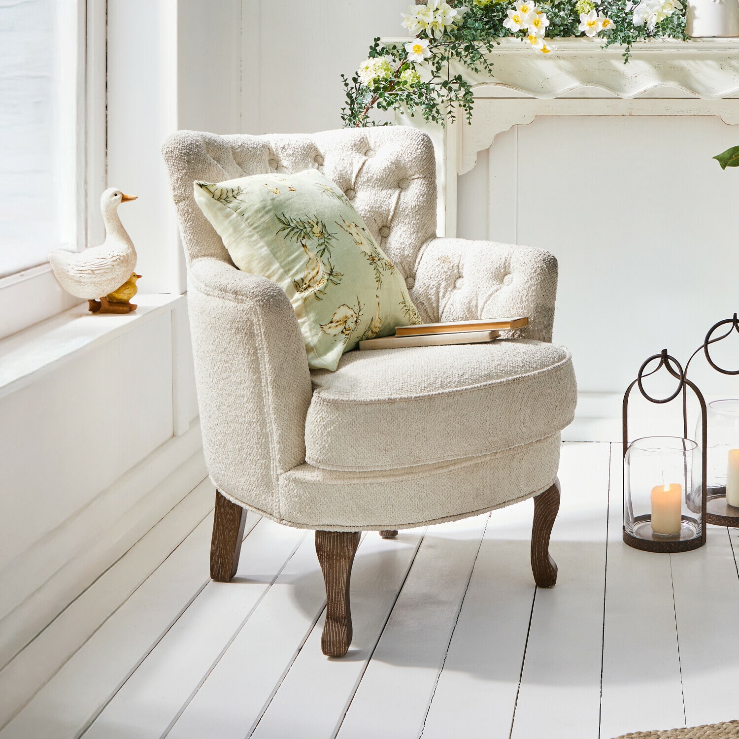 Poltrona Calgary Poltrona beige in tessuto morbido con gambe in legno e cuscino verde con fiori in ambiente luminoso