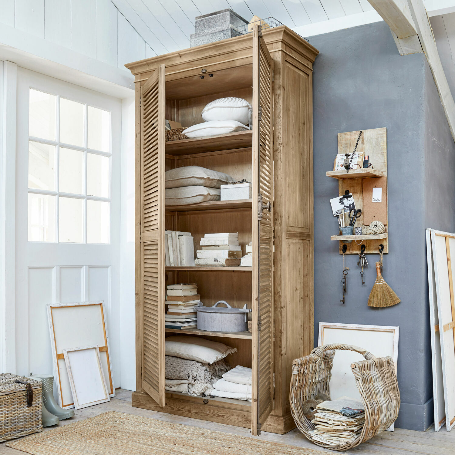 Armadio Farnham Armadio in legno con ante a persiana color naturale, decorato con cuscini e libri in ambiente luminoso