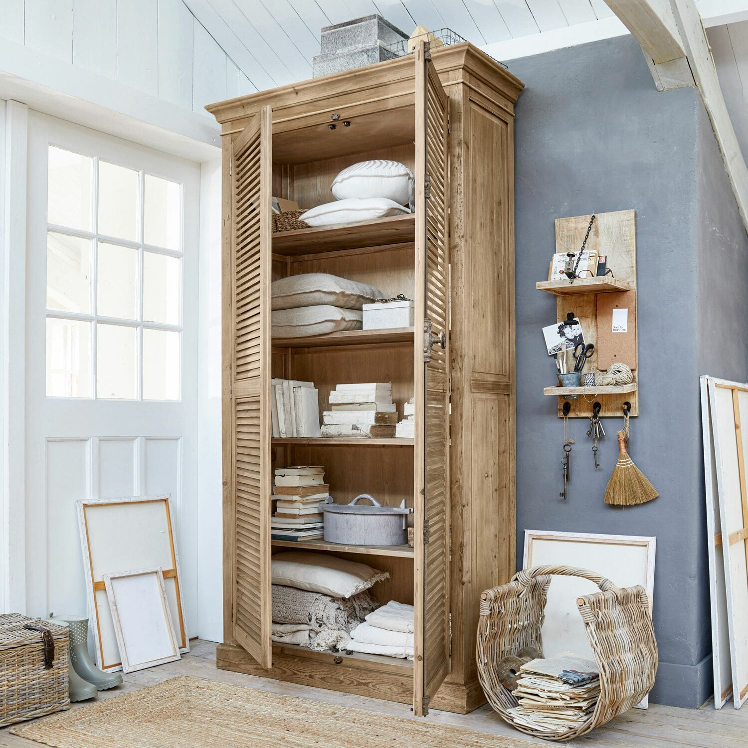 Armadio Farnham Armadio in legno con ante a persiana color naturale, decorato con cuscini e libri in ambiente luminoso