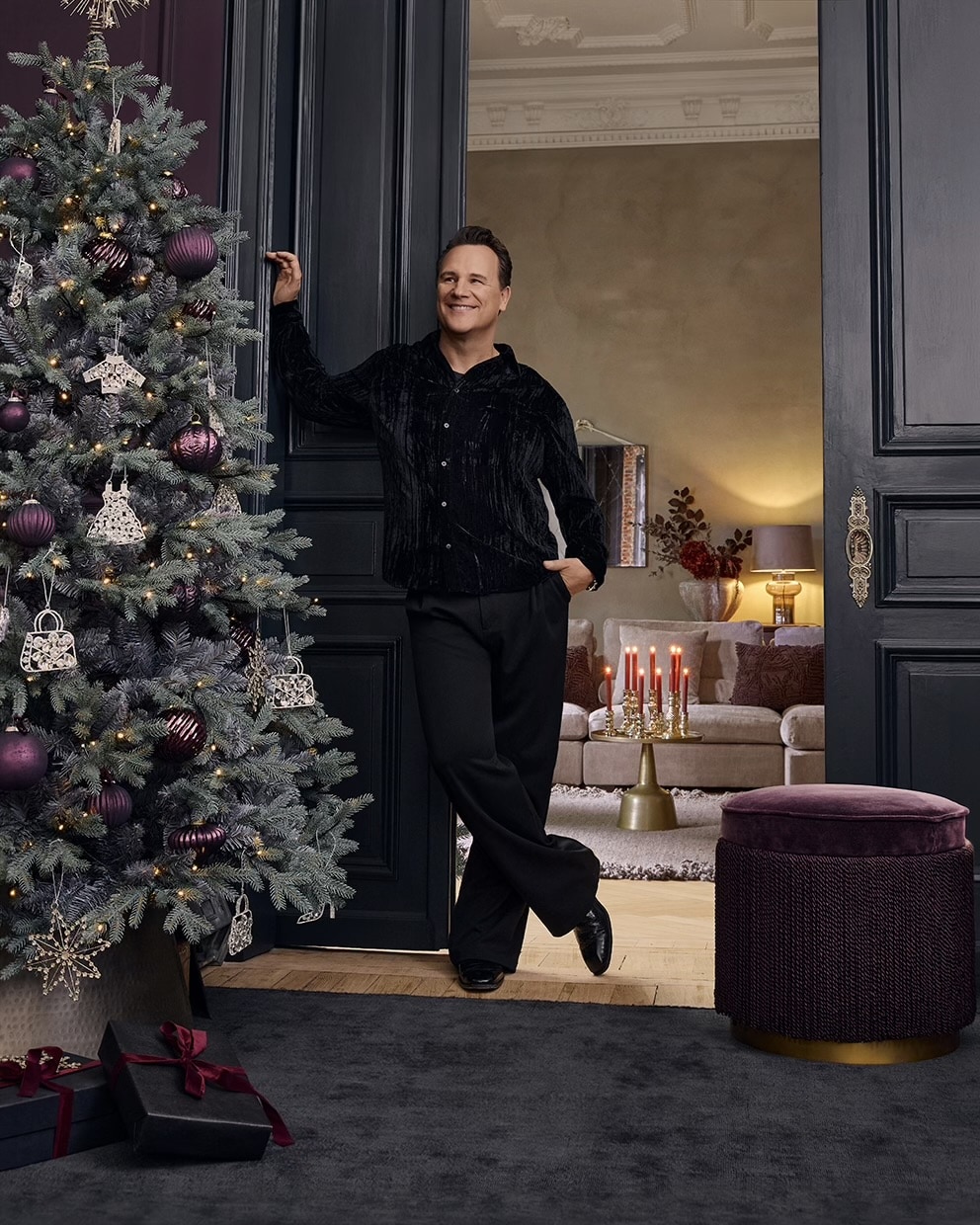Uomo con abbigliamento nero accanto ad albero di Natale decorato con palline viola in soggiorno elegante con candele rosse accese