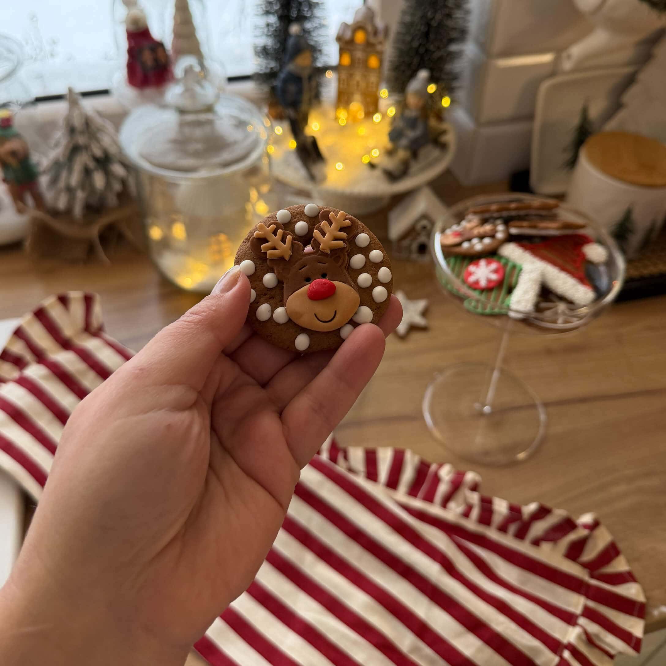 Mano che tiene un biscotto di pan di zenzero con renna e puntini bianchi, decorazioni natalizie sullo sfondo