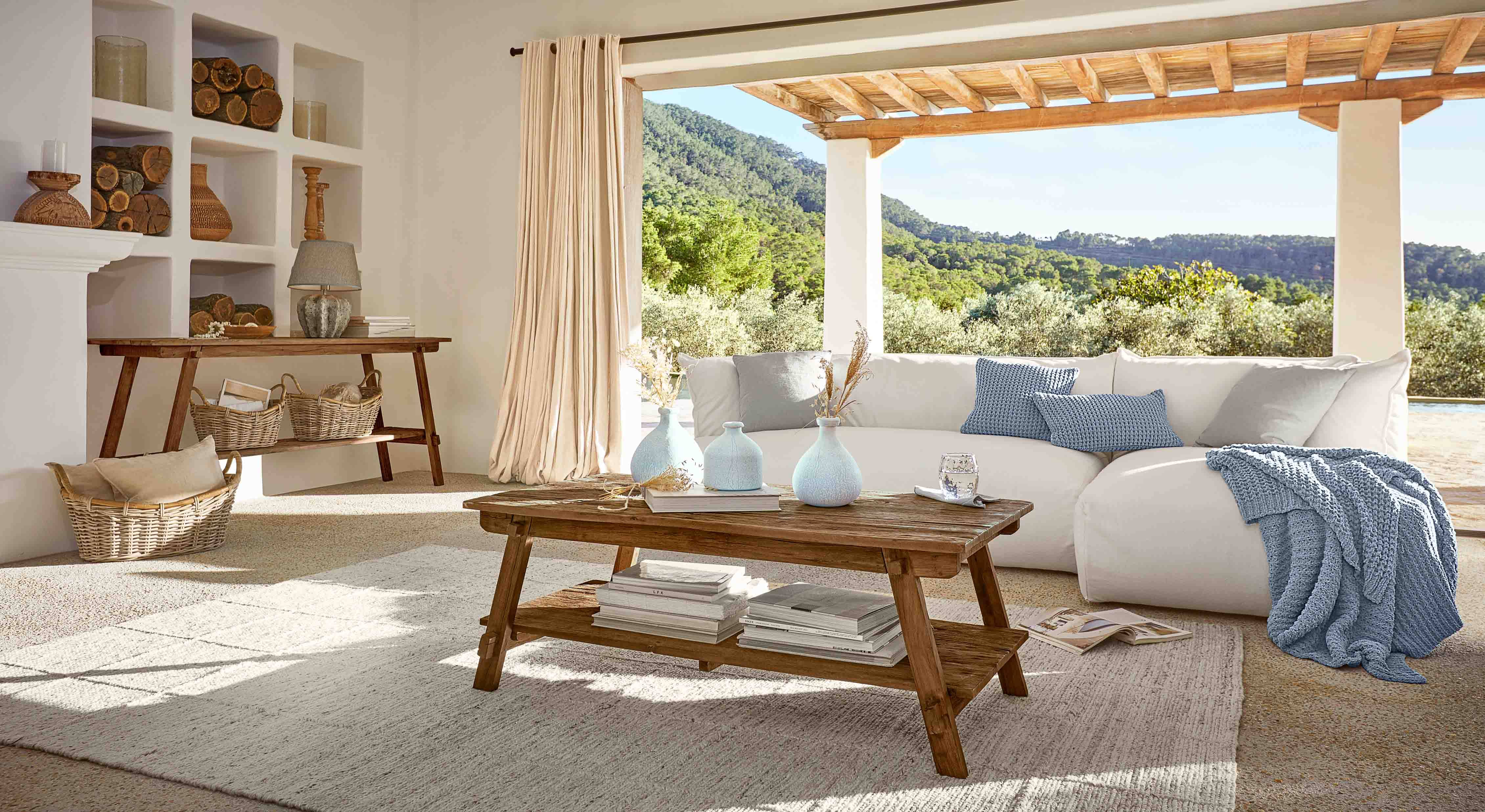 Soggiorno con tavolino in legno, divano bianco e coperta blu, decorazioni naturali e vista sul verde