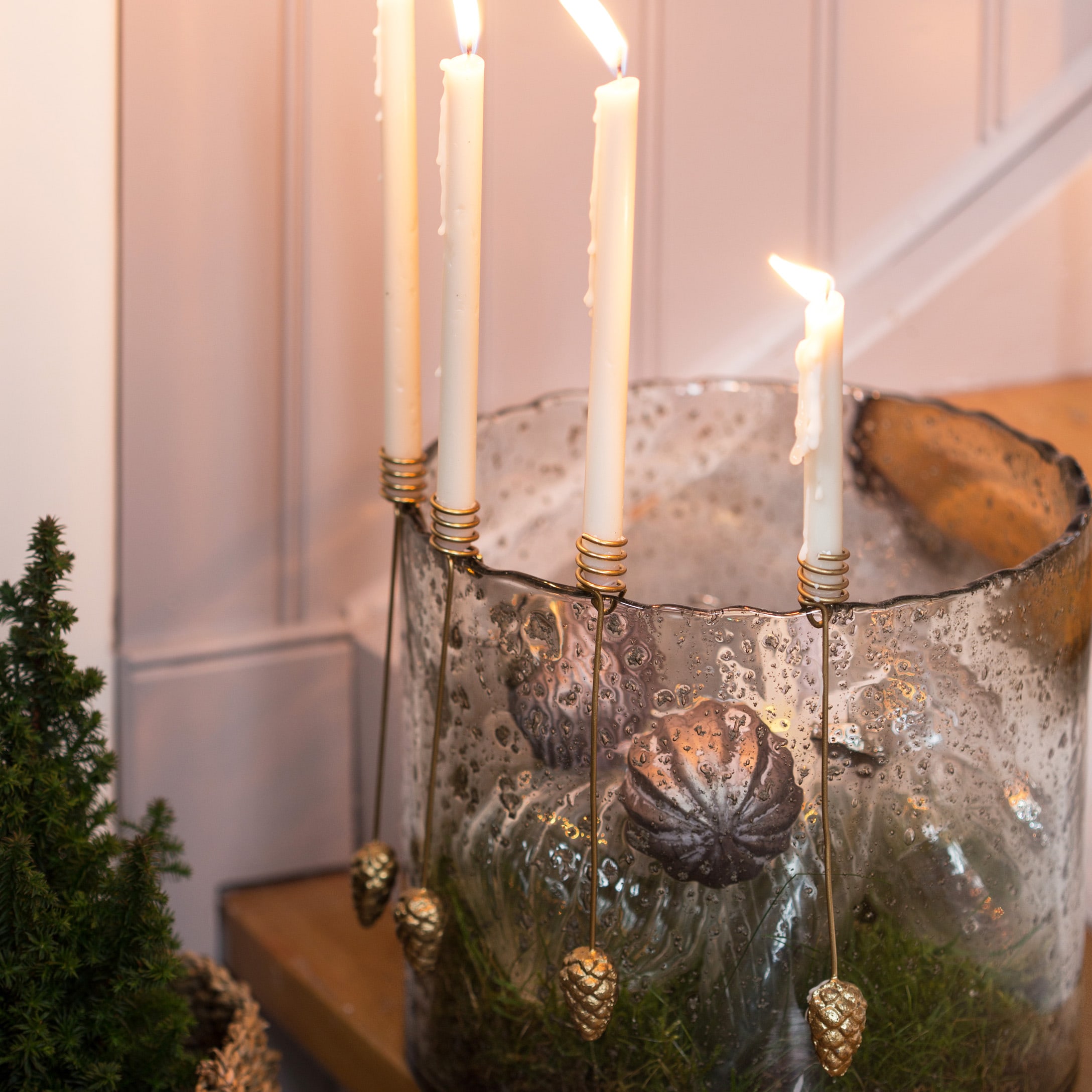Portacandele in vetro con candele bianche e decorazioni dorate a forma di pigne su tavolo in legno