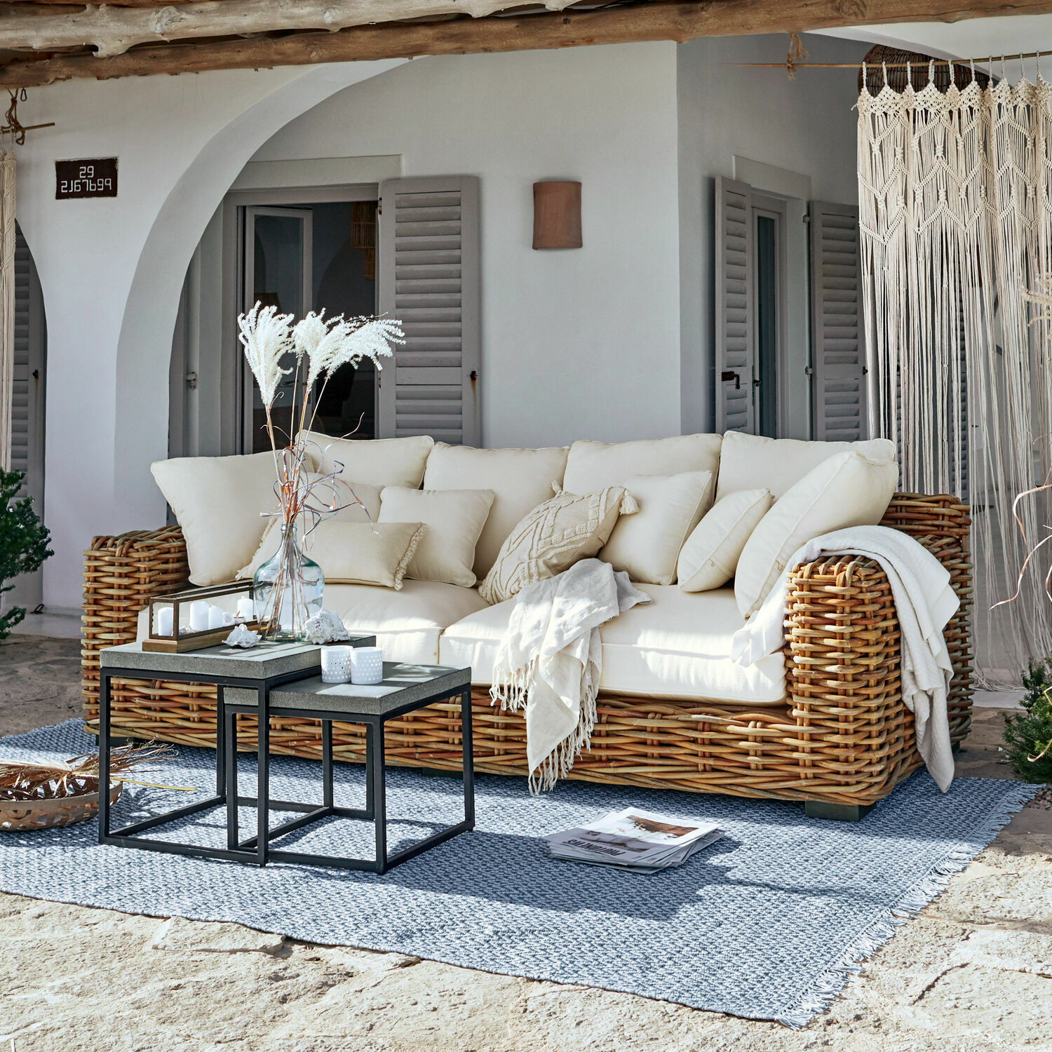 Sofà Rimini Divano in rattan con cuscini crema e coperta su tappeto blu in terrazza soleggiata