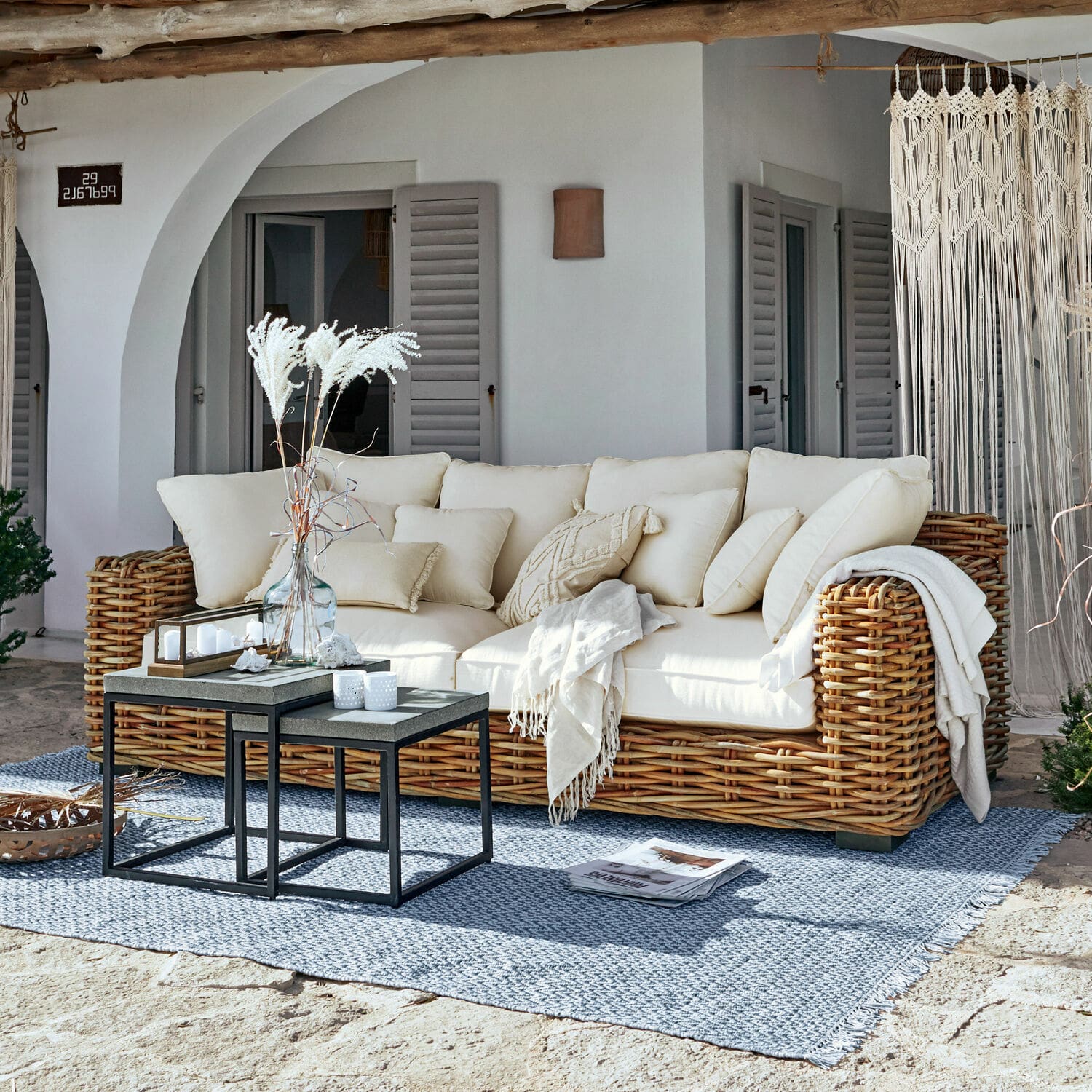 Sofà Rimini Divano in rattan con cuscini crema e coperta su tappeto blu in terrazza soleggiata