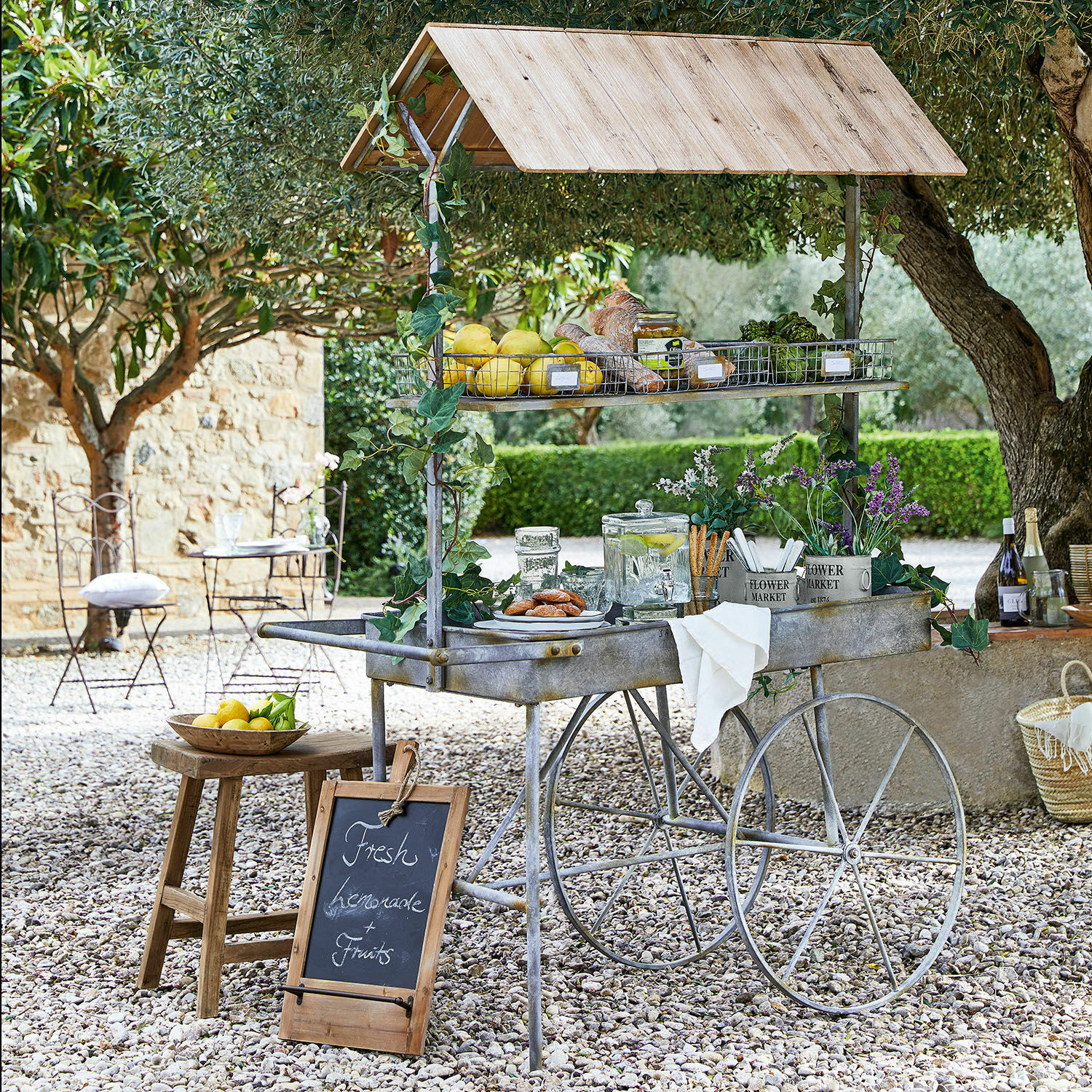 Tavolo per piante Granby Carretto in metallo con tetto in legno, frutta fresca e limonata in un giardino soleggiato