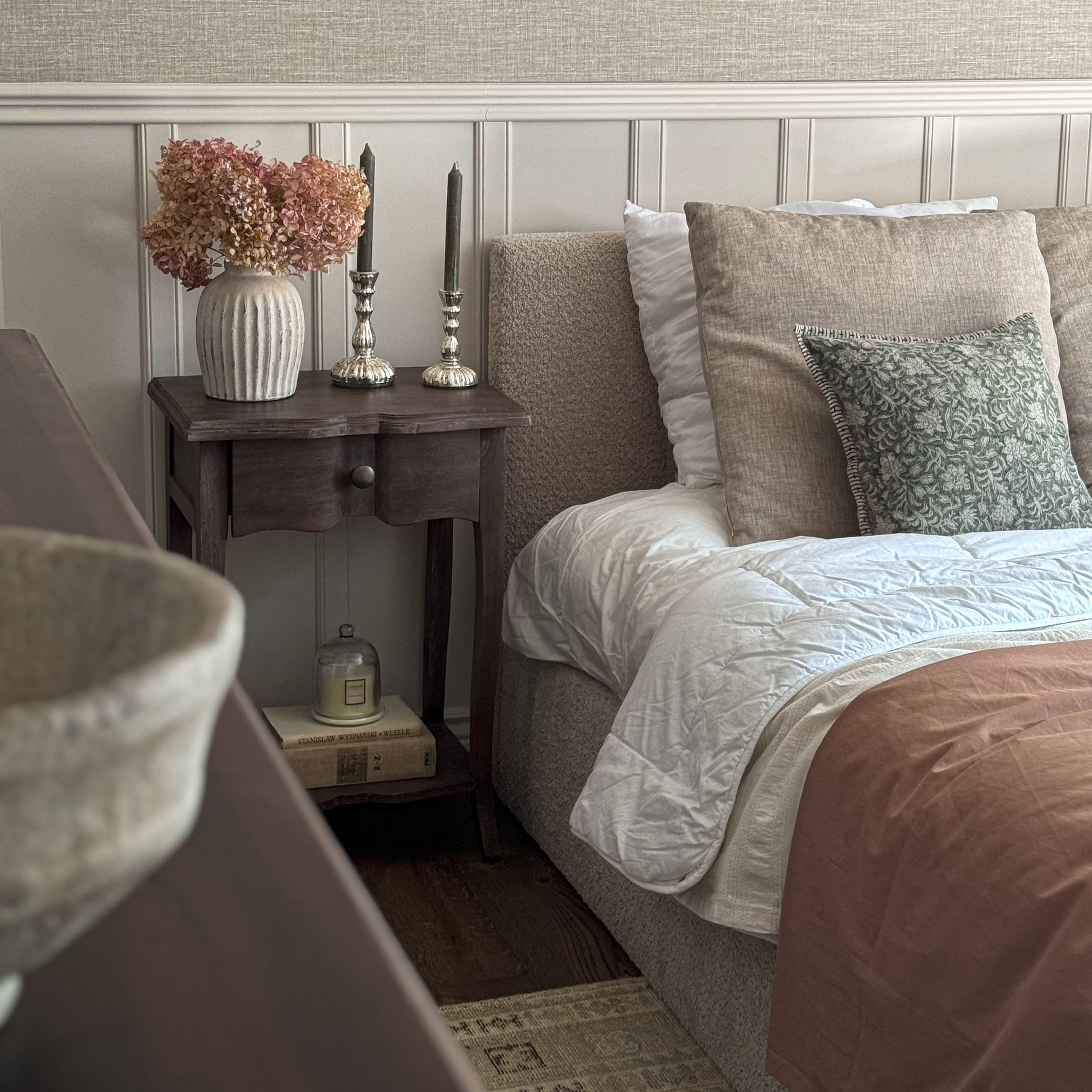 Camera da letto accogliente con biancheria beige, comodino in legno e vaso decorativo con fiori secchi