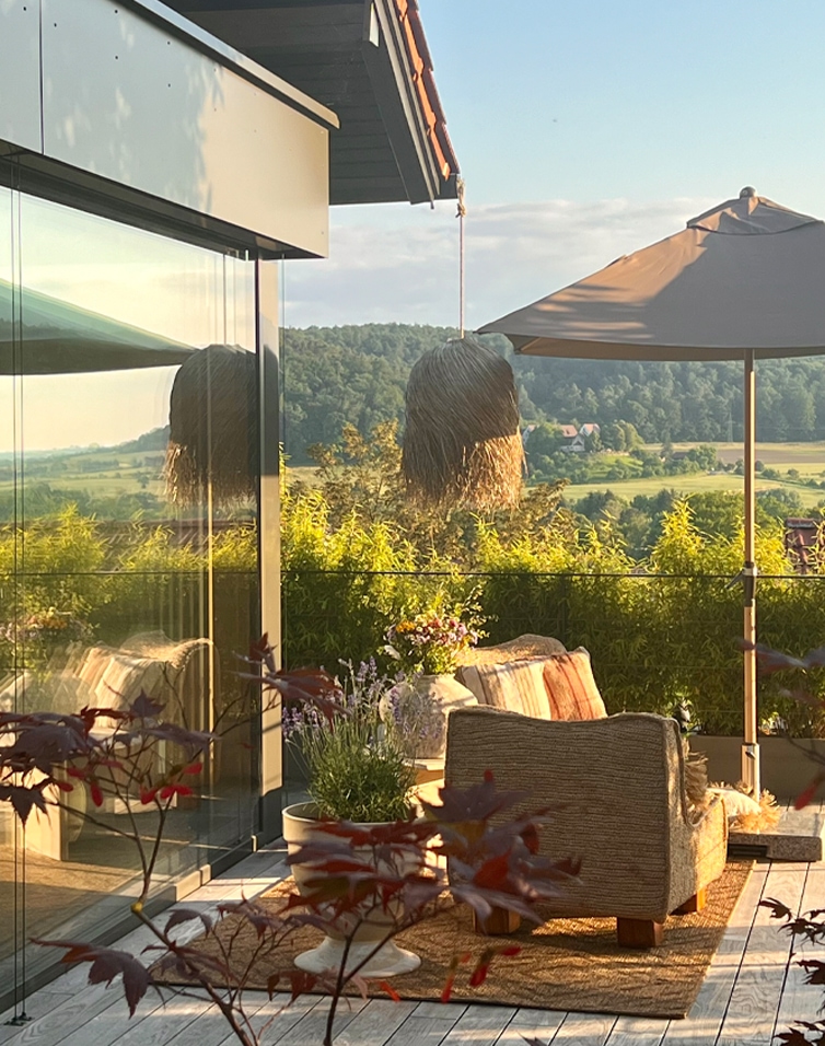 Accogliente terrazza con divano beige, ombrellone e piante davanti a colline verdi in una giornata di sole