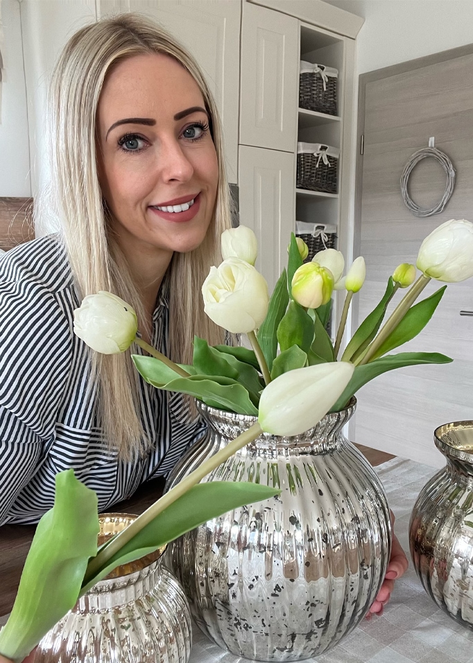 Donna con capelli biondi accanto a vaso argentato con tulipani bianchi in interno moderno