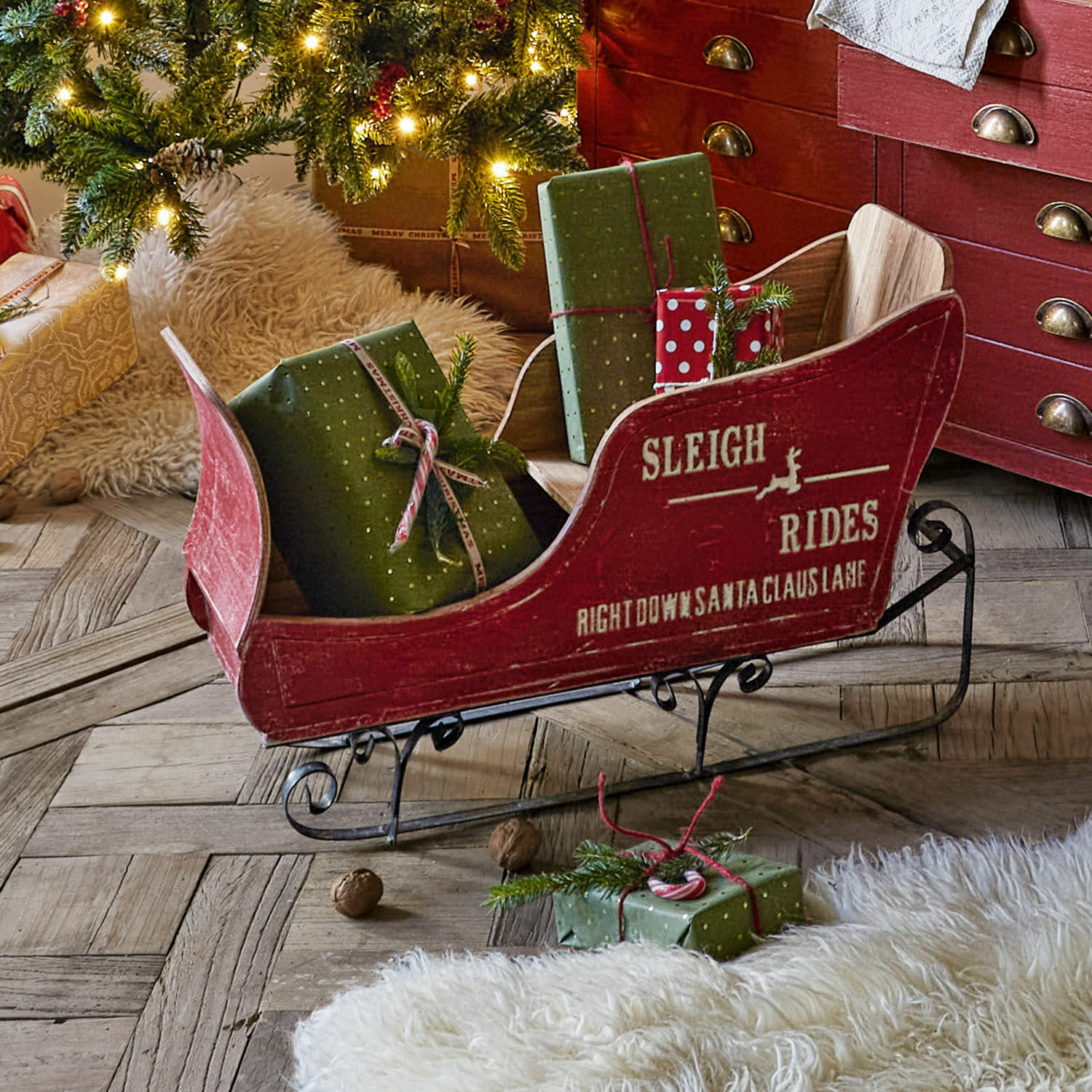 Slittino decorativo Maélys Slitta in legno rossa con pattini in metallo e regali verdi su pavimento in legno vicino all'albero di Natale