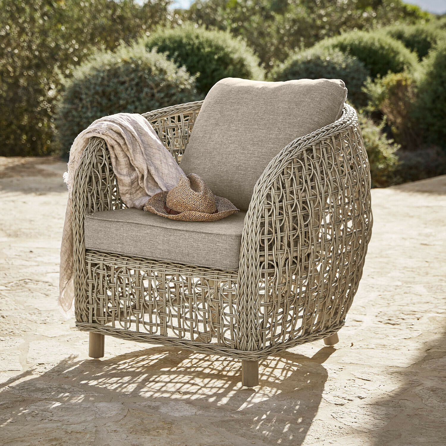 Poltrona da giardino in vimini beige con cuscini beige chiaro, cappello di paglia e coperta appoggiati