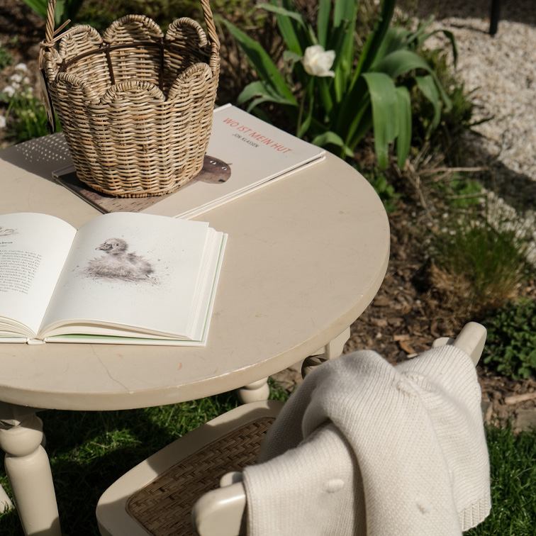 Tavolo rotondo beige da giardino con cesta in vimini e libri aperti sull'erba al sole