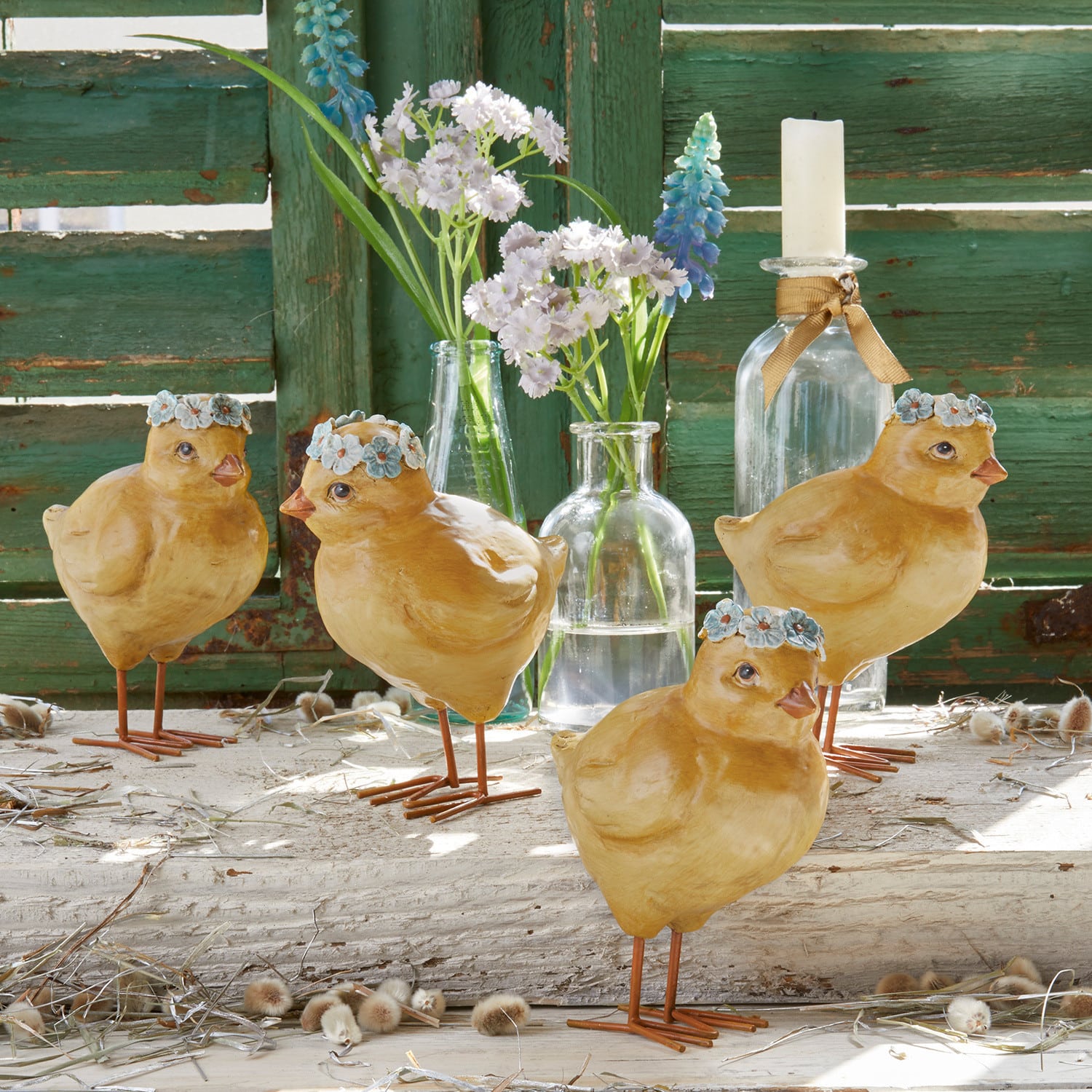 Set di 4 pulcini Dorette Quattro pulcini decorativi gialli in materiale con corone di fiori blu davanti a una parete di legno verde