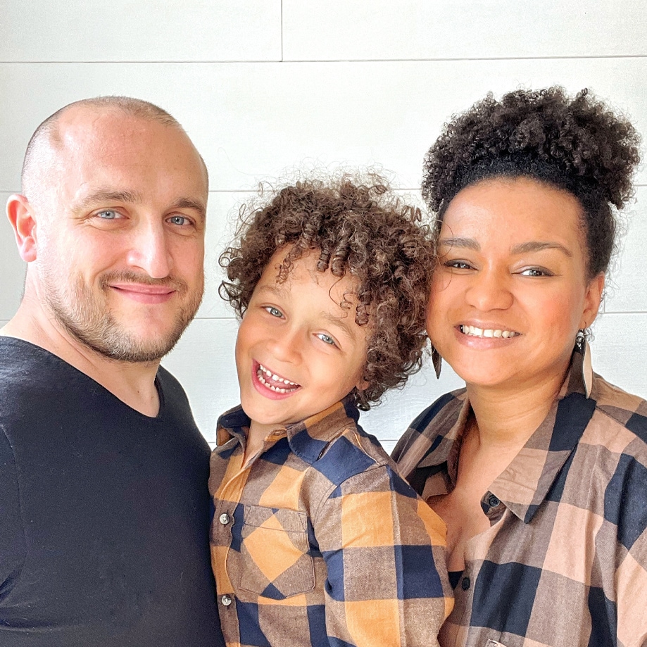 Famiglia sorridente davanti a parete chiara, bambino e adulti in abbigliamento casual.