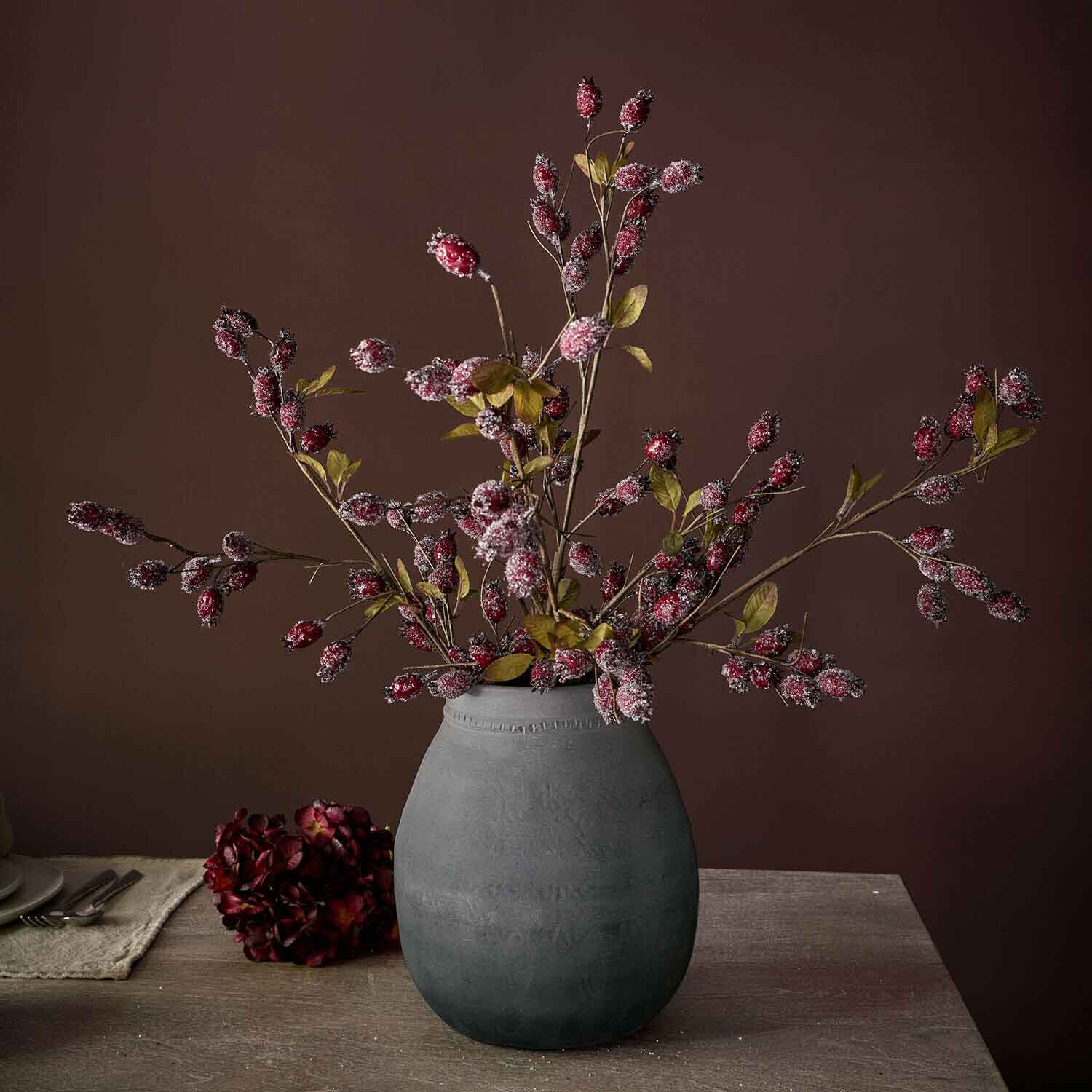 Vaso in ceramica grigia con rami decorativi con bacche rosse e foglie verdi su tavolo in legno