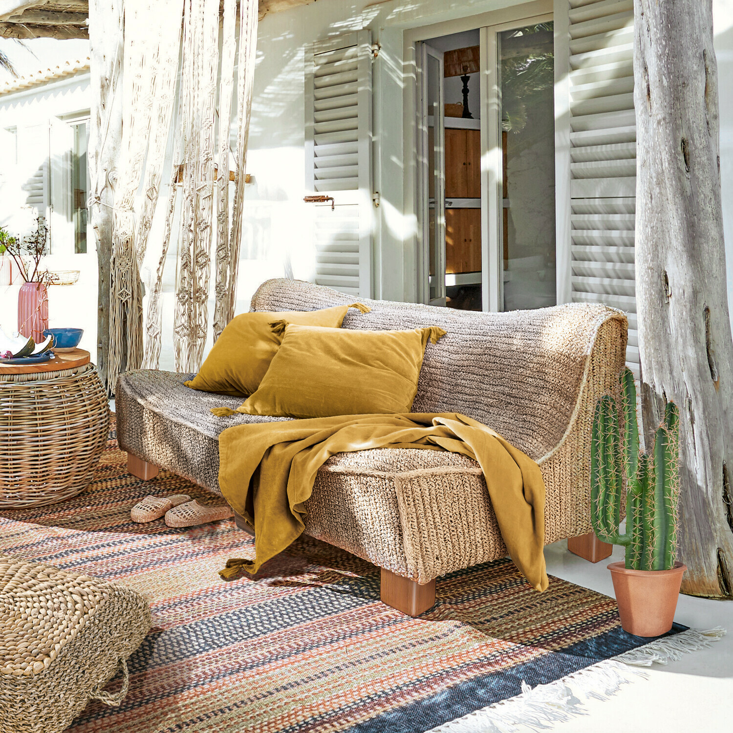 Divano Bocage Divano in materiale intrecciato colore naturale con cuscini e coperta senape su terrazza soleggiata
