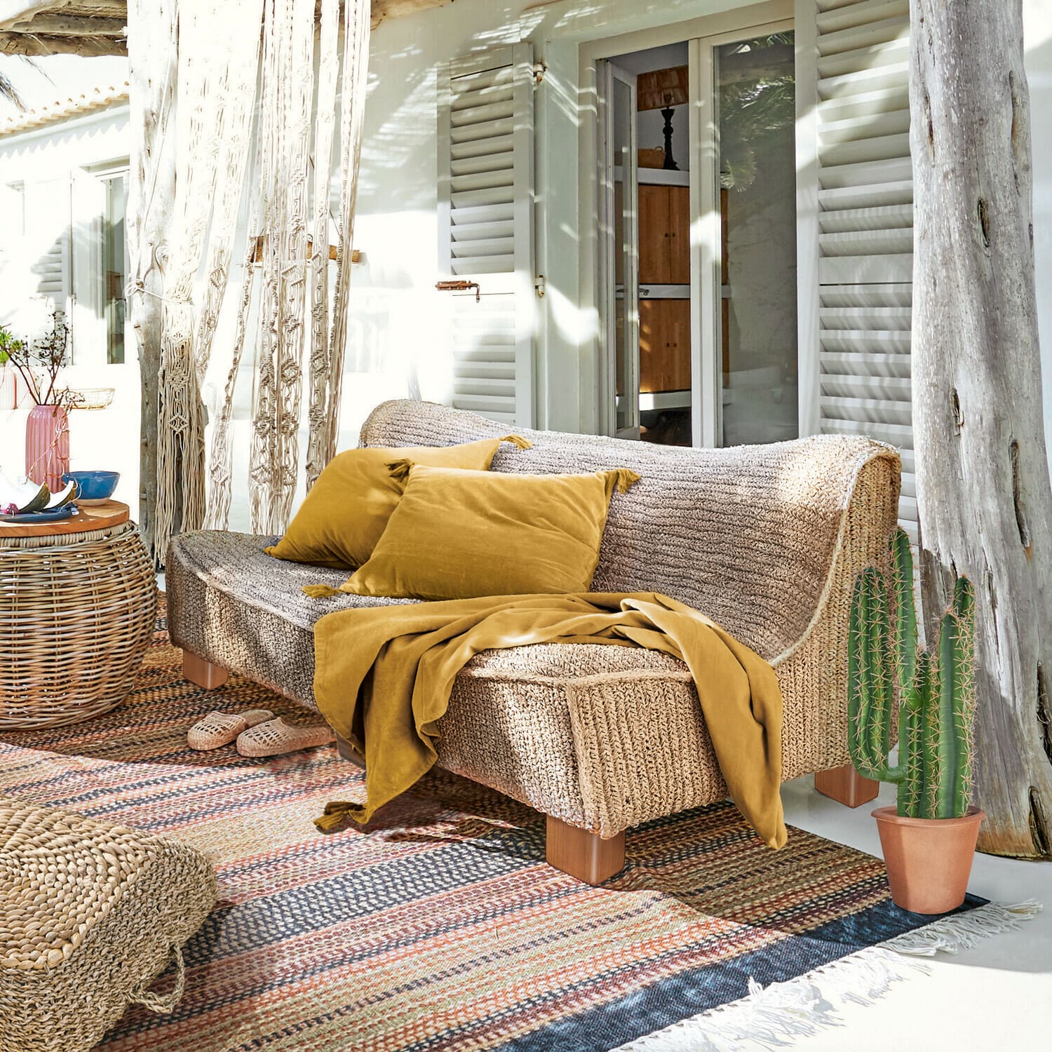 Divano Bocage Divano in materiale intrecciato colore naturale con cuscini e coperta senape su terrazza soleggiata