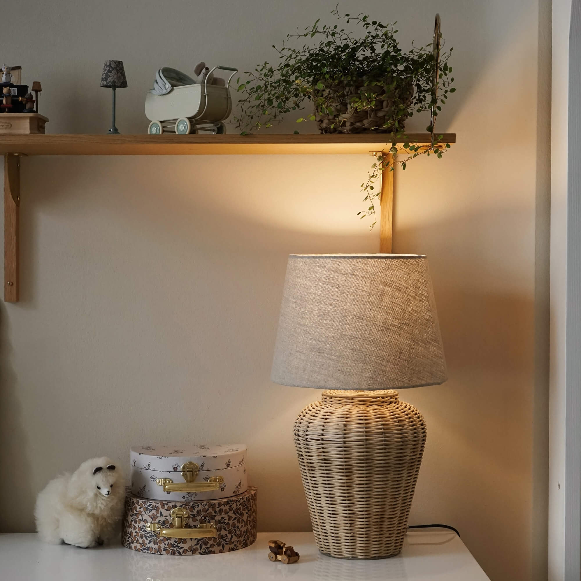 Lampada da tavolo in vimini con paralume beige su tavolo bianco con decorazioni e pianta su mensola
