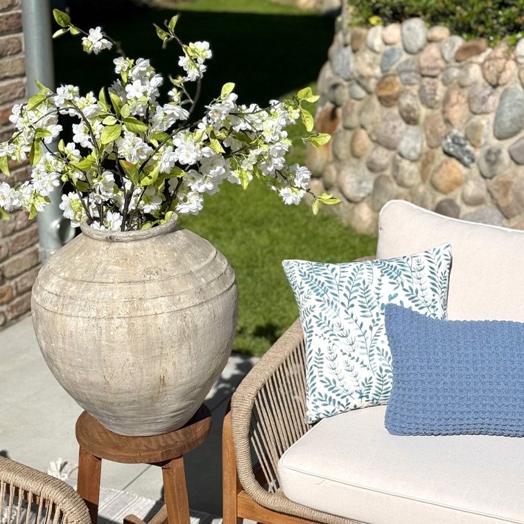 Grande vaso in ceramica beige con fiori bianchi su supporto in legno accanto a divano con cuscini in giardino