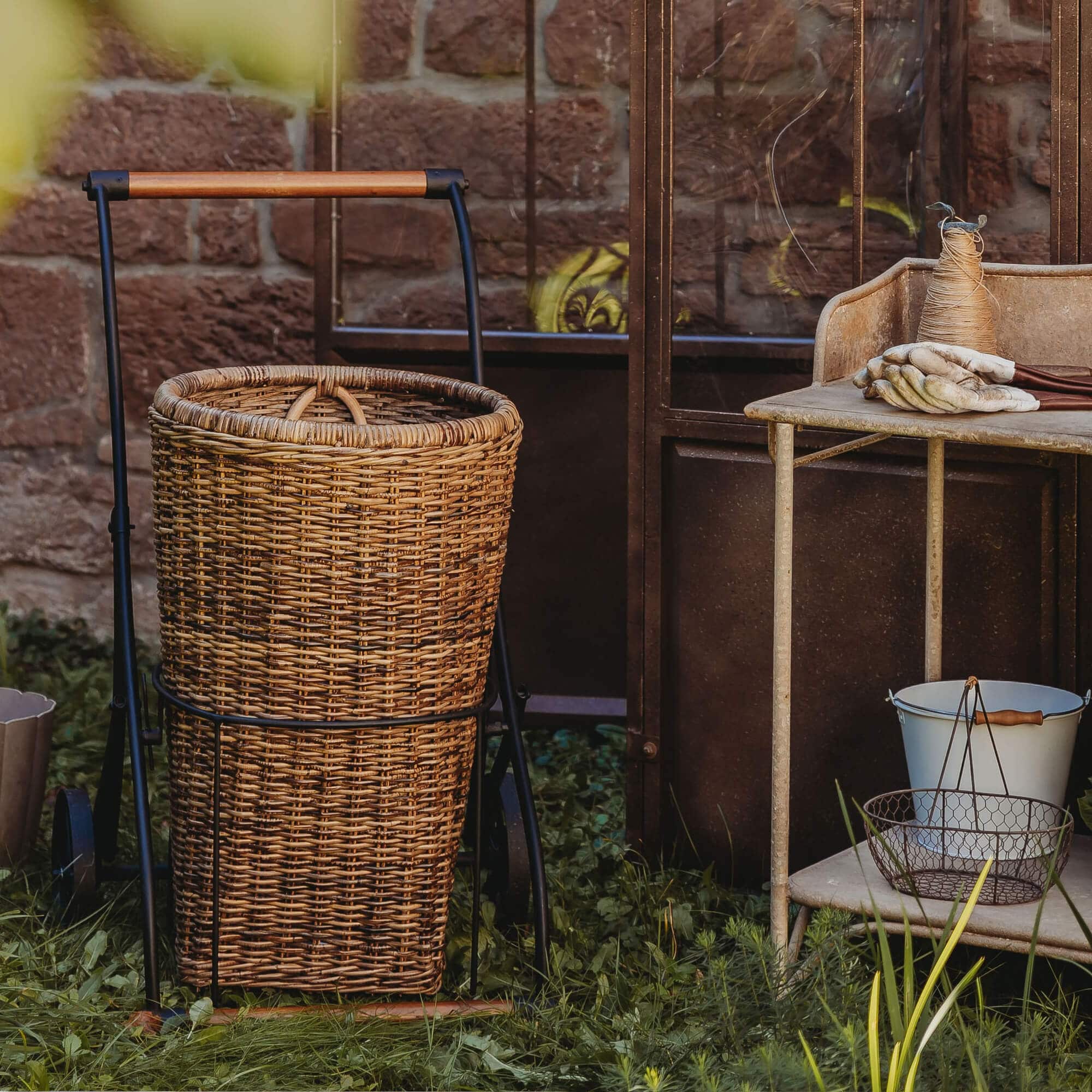 Carrello con cesta in vimini e struttura metallica sull'erba vicino a muro di mattoni, attrezzi da giardino accanto