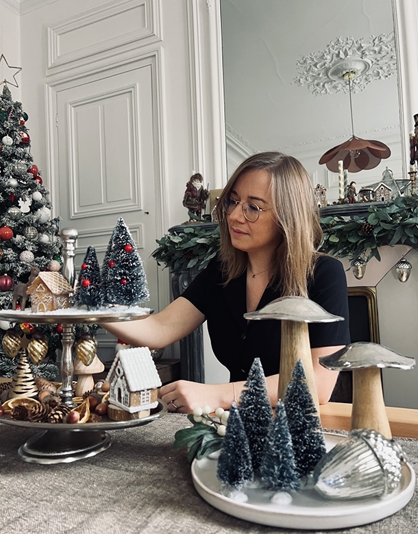 Donna che decora tavolo natalizio con piccoli alberi e casette in un ambiente accogliente.