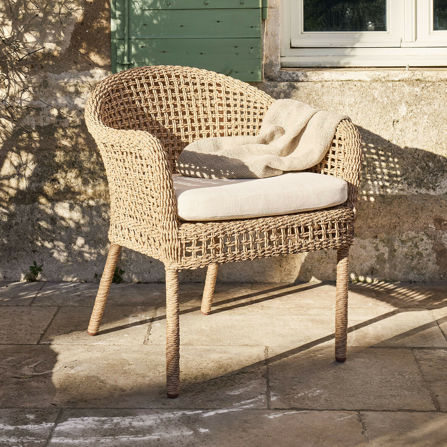 Poltrona in rattan intrecciato naturale con cuscino chiaro e coperta beige su terrazza in pietra